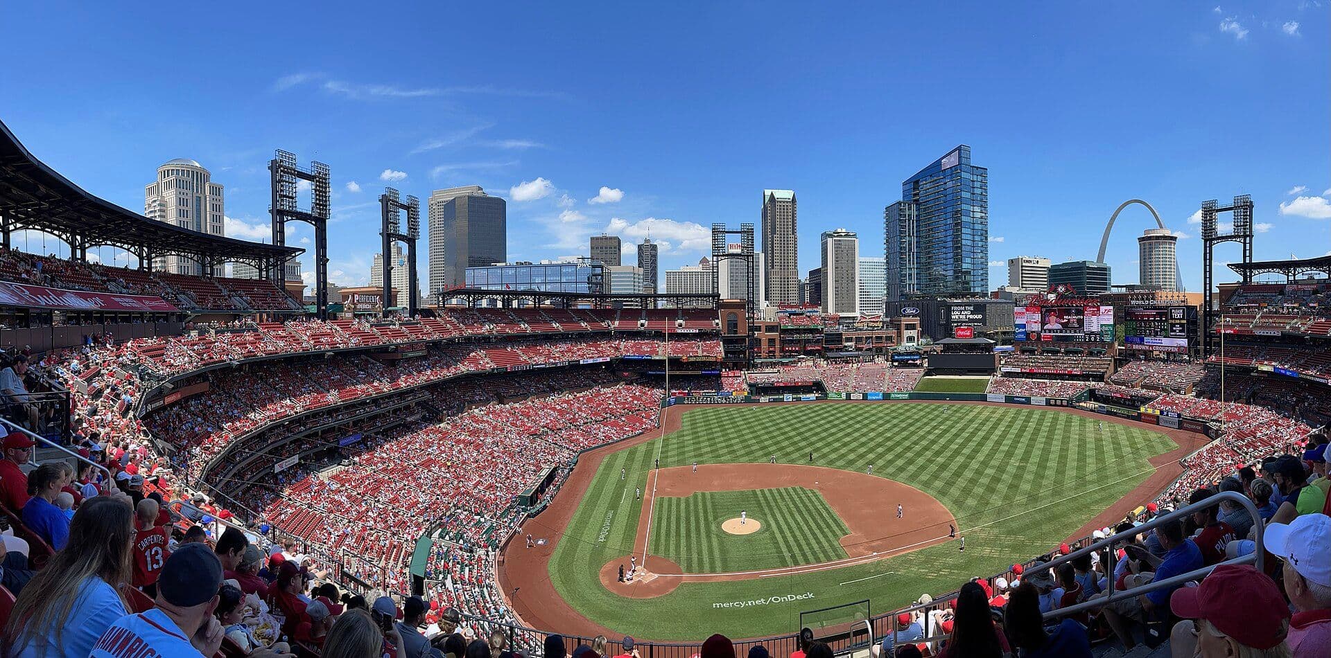 Wide panoramic view of Busch Stadium in St. Louis during a day game, with the field, packed stands and the downtown skyline and Gateway Arch behind the outfield.